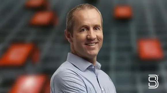 A man in a light blue shirt smiles while standing in front of a blurred background with red rectangles and a white logo in the bottom right corner.