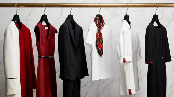 A row of formal and semi-formal women's clothing, including dresses, blazers, shirts, and a scarf, hangs on a gold rack against a marble wall.