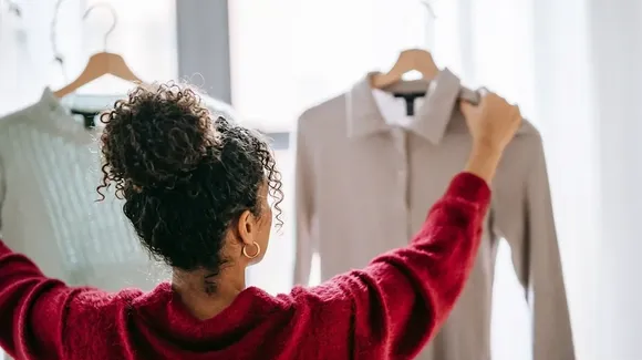 A person with curly hair in a red sweater holds up two shirts on hangers, comparing them in front of a window.