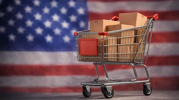 A shopping cart filled with cardboard boxes stands in front of a blurred American flag backdrop.