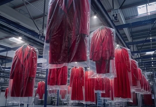 Rows of red garments in protective plastic covers hanging from a ceiling conveyor system in a warehouse.
