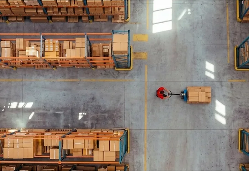 An overhead view of a warehouse worker using a pallet jack to move a box through an aisle between tall storage racks filled with cardboard boxes.
