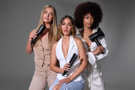 Three women posing with hair styling tools against a gray background. One holds a flat iron while the other two hold hair dryers.