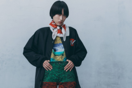 Person with short black hair wearing a black jacket, colorful patterned shorts, and a red and white scarf, standing against a light-colored wall.