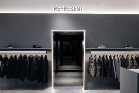 Clothing display in a modern store featuring jackets and shoes. A central, illuminated doorway leads to another area, with "REPRESENT" signage above.