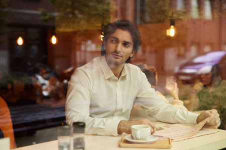 A person in a white shirt is sitting at a cafe table, holding a newspaper. A cup of coffee is in front of them. Reflections of street and lights are visible on the window.