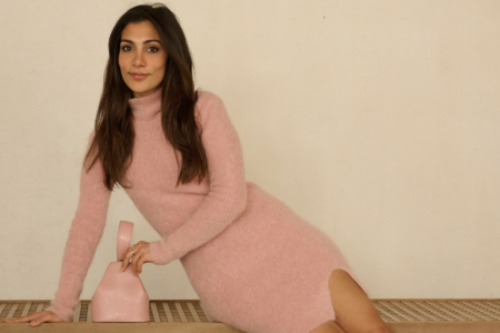 A woman in a light pink long-sleeve dress sits on a bench, with her hand resting on a small pink handbag against a plain background.