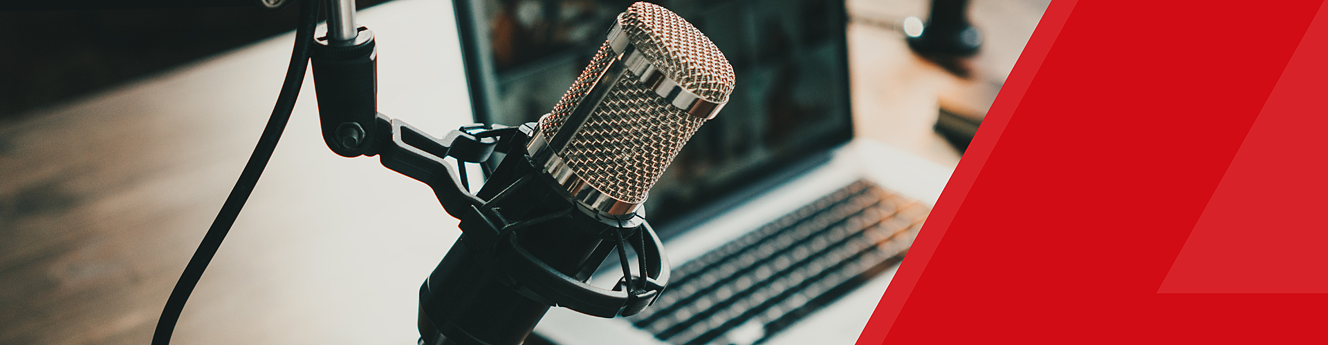 A close-up of a microphone in front of an open laptop on a wooden desk, with a red diagonal graphic on the right side.