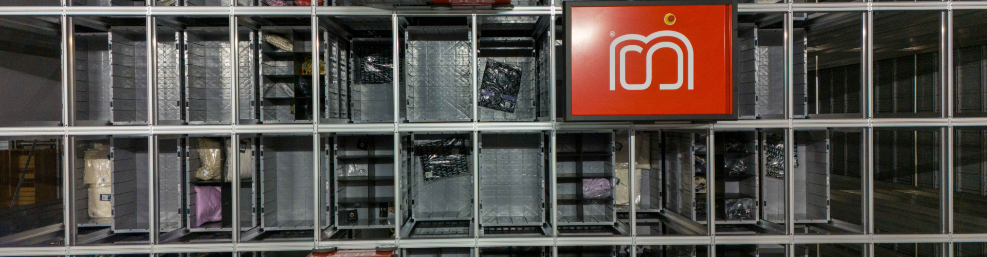 Top-down view of a warehouse shelving system with red panels featuring a white logo, containing organized products in a grid-like structure.