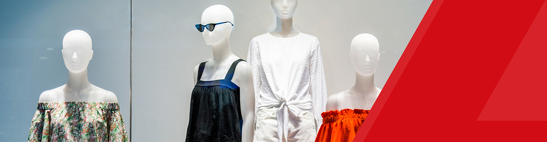 Mannequins displaying various dresses and a pair of sunglasses in a store window, with a red geometric design on the right side.