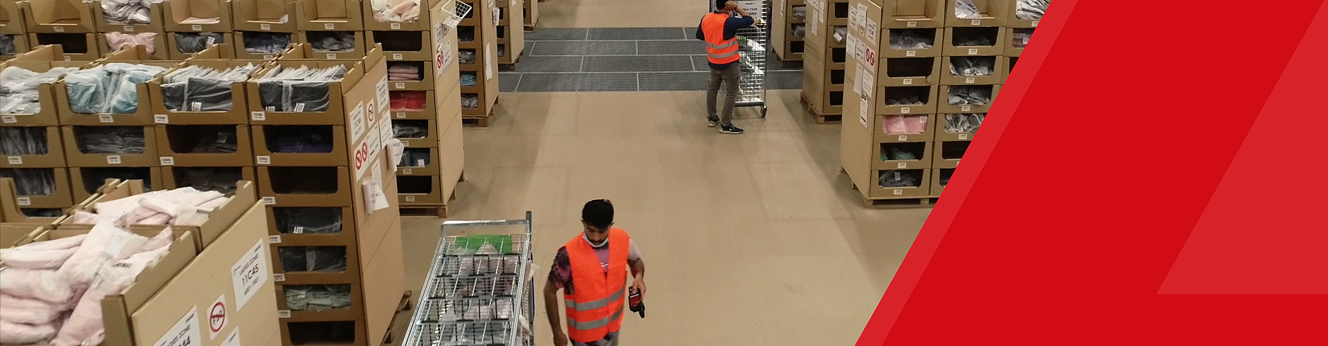 Workers in orange vests organize inventory in a warehouse aisle, surrounded by shelves filled with boxes and products.