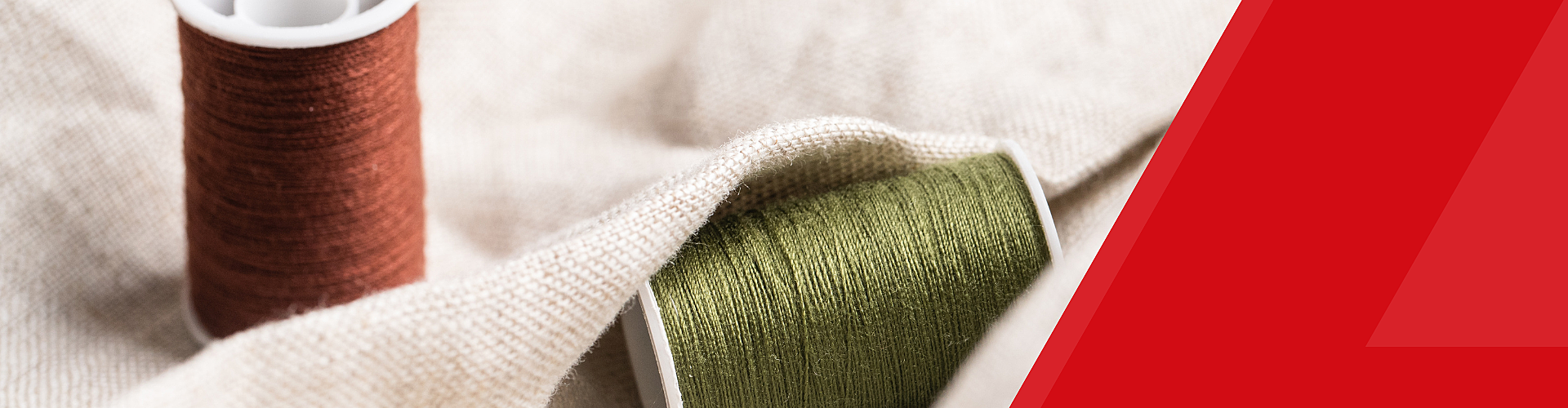 Two spools of thread, one red and one green, rest on a beige fabric. A red geometric shape is on the right edge of the image.