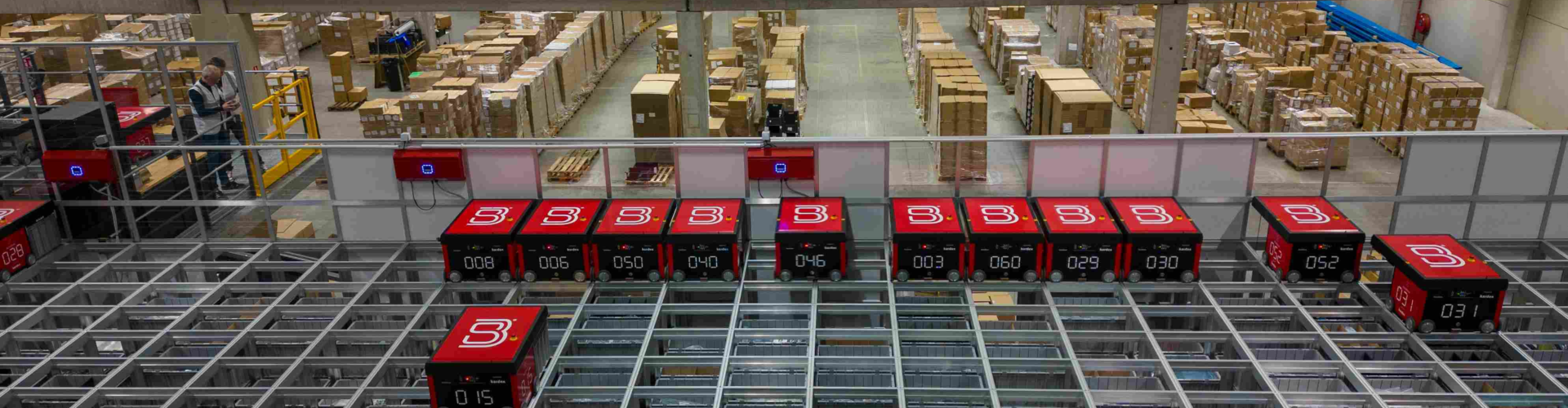 Warehouse interior with automated storage and retrieval system. Red robotic carts move on a grid above shelves filled with boxes, illustrating modern logistics and inventory management.