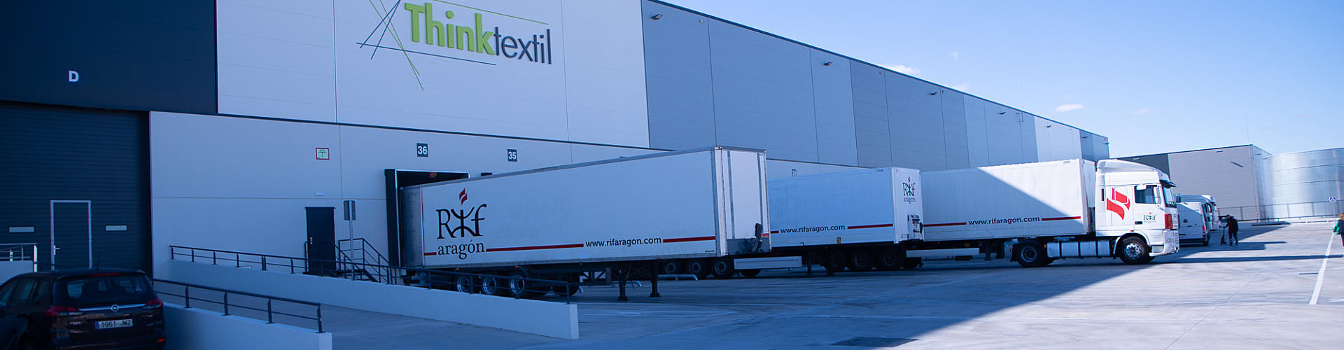Three large white trucks parked outside a modern industrial building with "Think Textil" signage. Clear blue sky.