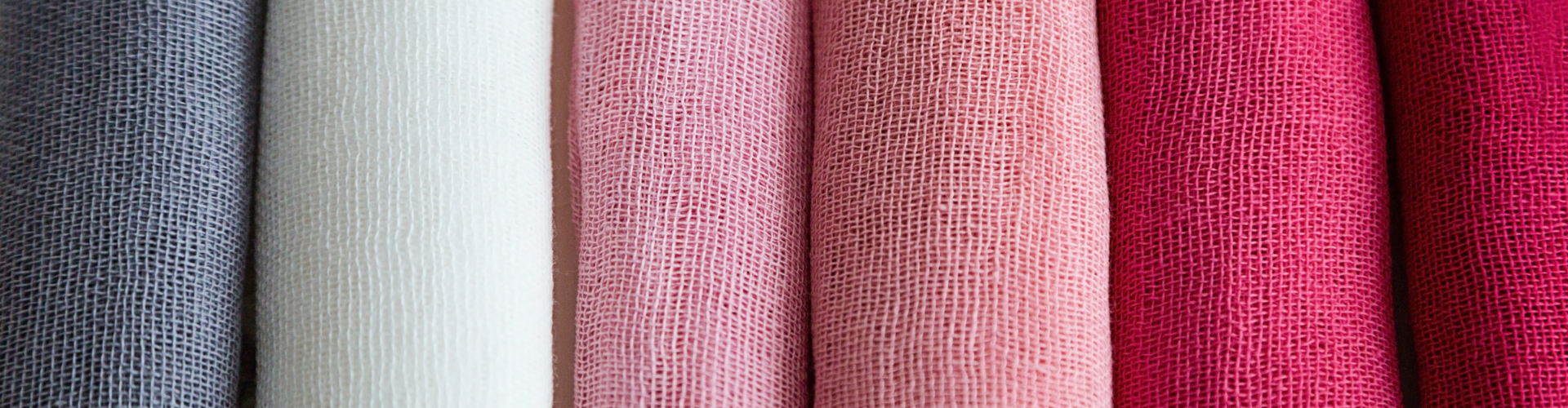 Close-up of six rolls of fabric in various colors: gray, off-white, light pink, pink, dark pink, and red, arranged vertically side by side.
