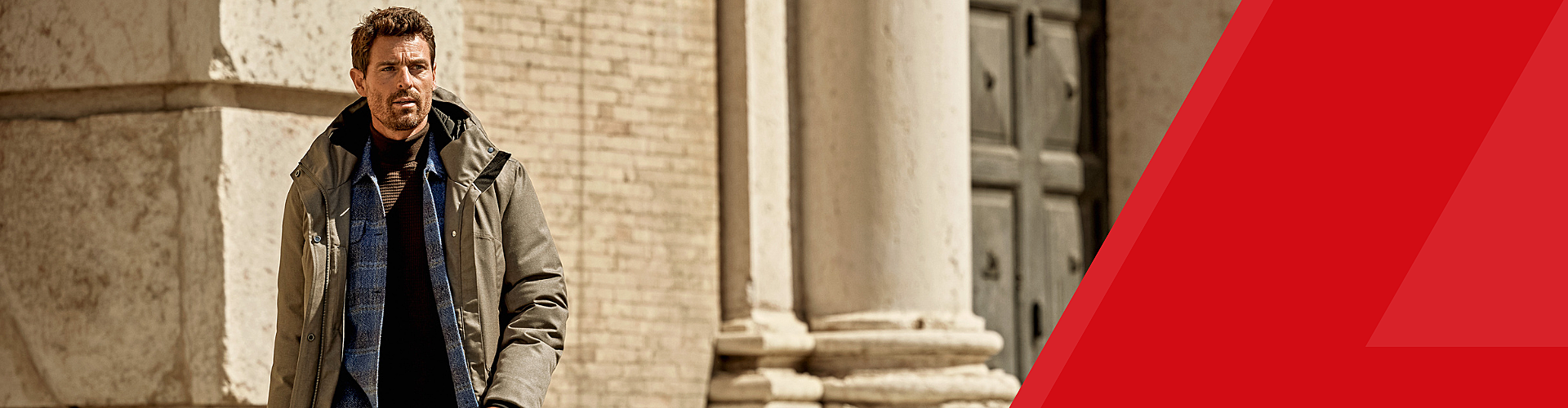 A man wearing a gray jacket and blue sweater stands outdoors near large stone columns and a brick wall, with a red geometric shape on the right.