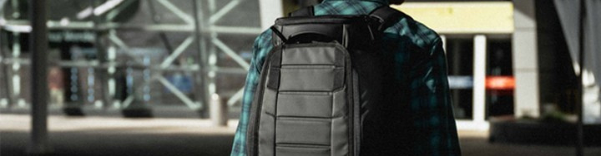 A person wearing a black beanie and plaid shirt stands outside a modern glass building with a large black backpack.