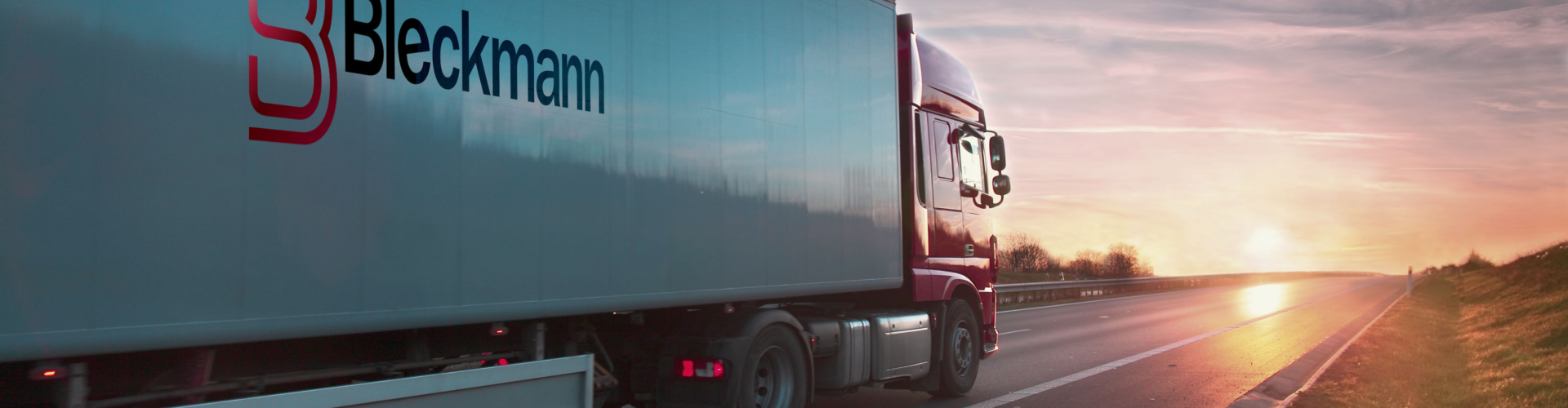 A Bleckmann-branded semi-truck drives on an empty highway at sunset, with the sky displaying clouds and contrails.