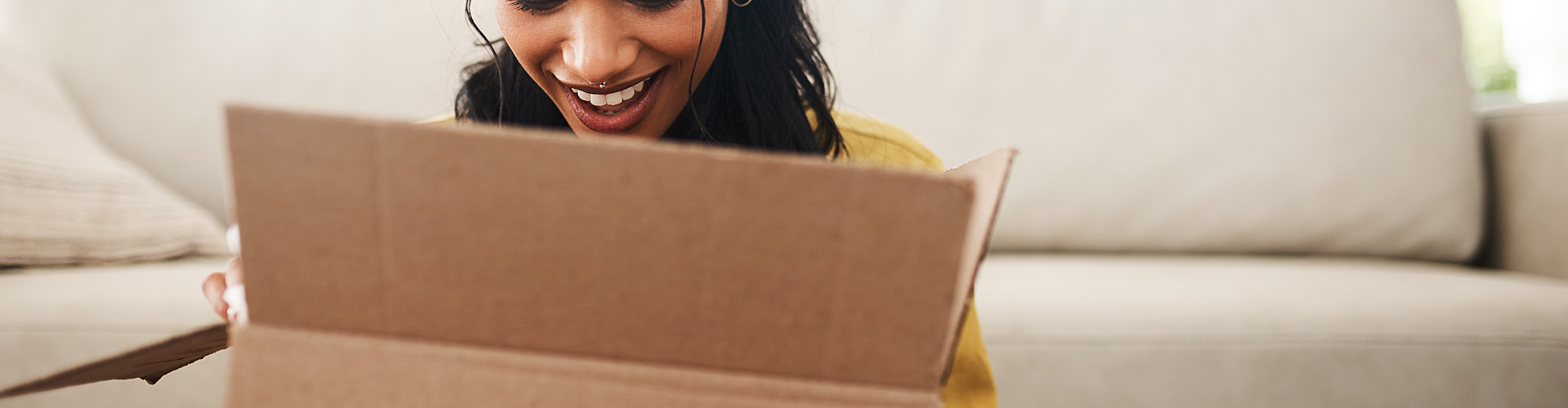 A woman sitting indoors opens a cardboard box and smiles while looking inside. A beige sofa is in the background.