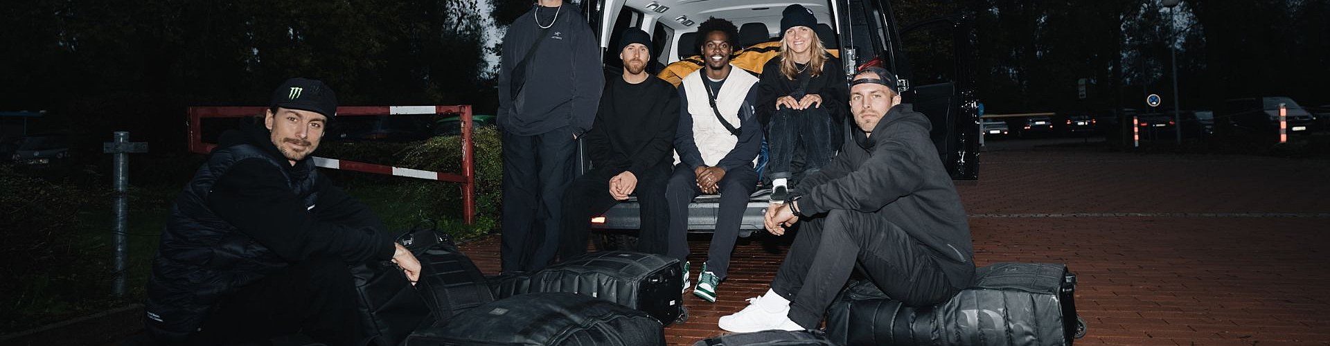 Six people sit and kneel around several black bags next to an open van in an outdoor setting with trees and brick pavement.