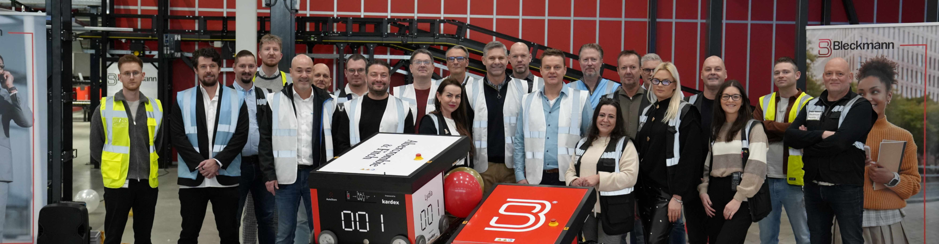 A group of people poses indoors at an industrial facility with two large numbered cubes in front, surrounded by red, black, white, and gold balloons.