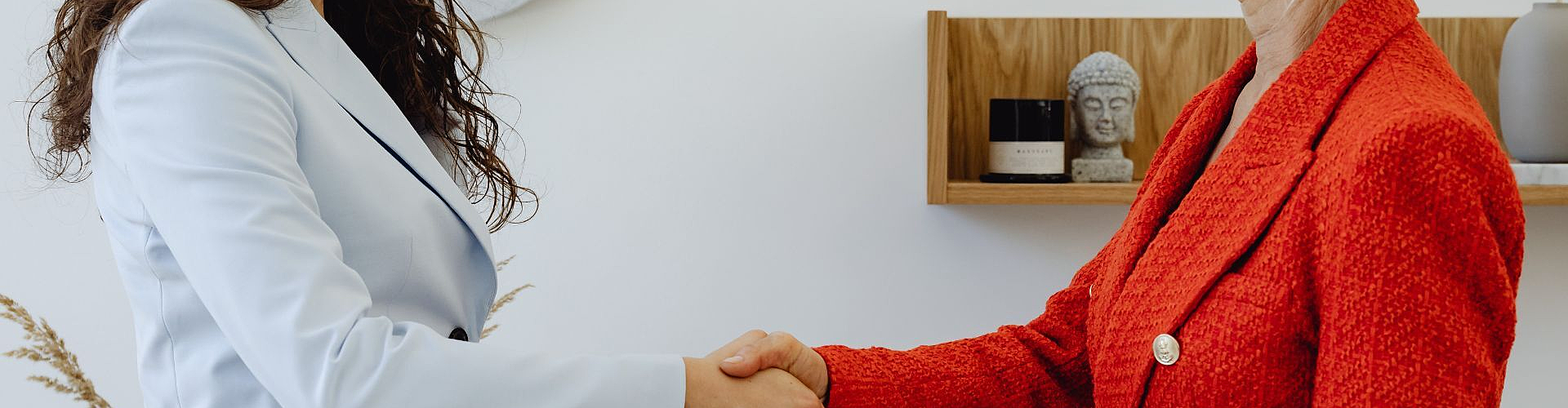 Two women in business attire are shaking hands in an office setting, standing in front of a shelf with a Buddha statue and decorative items.