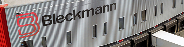 A gray industrial building with the Bleckmann logo and name on its exterior, above several loading dock doors.