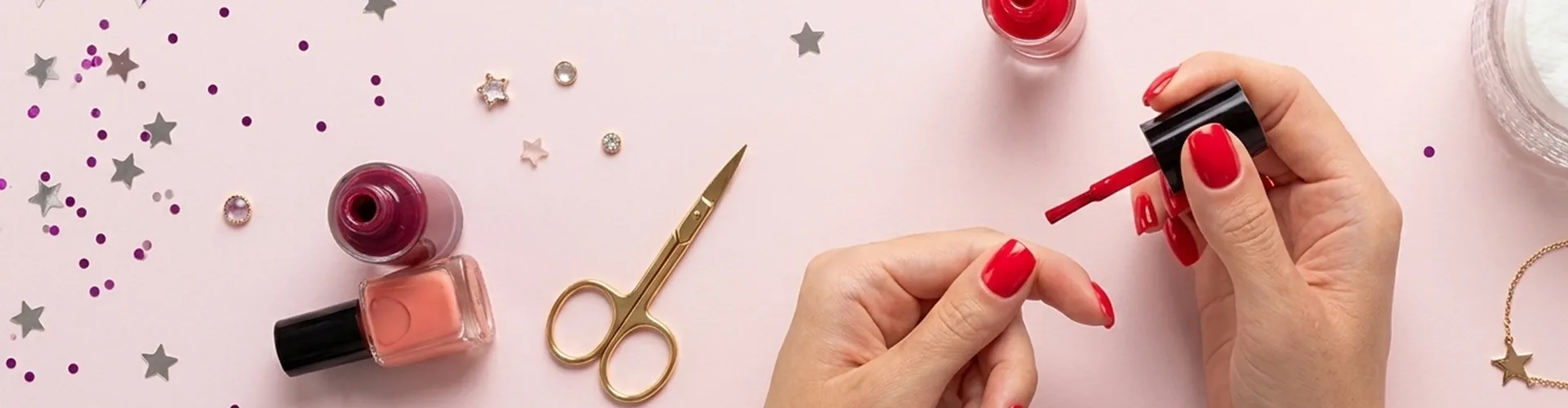 A person applies red nail polish to their nails on a table with nail polish bottles, scissors, confetti, and jewelry.