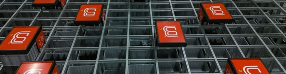 A warehouse automation system features red robotic units moving along a grid structure above storage bins.