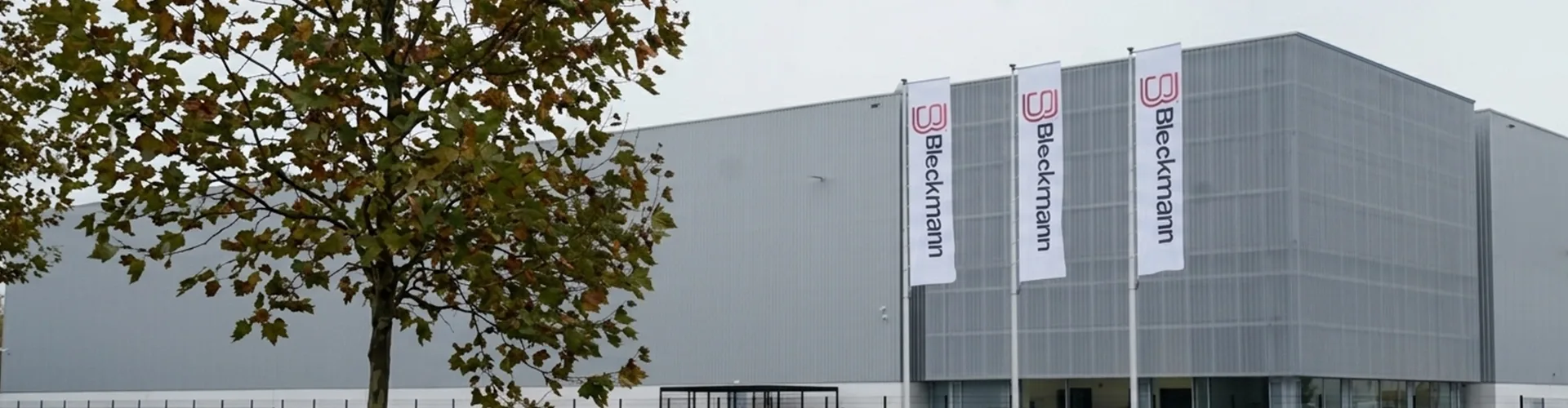 A large modern warehouse building with two vertical flags displaying the name "Blickmann" and a tree in the foreground.