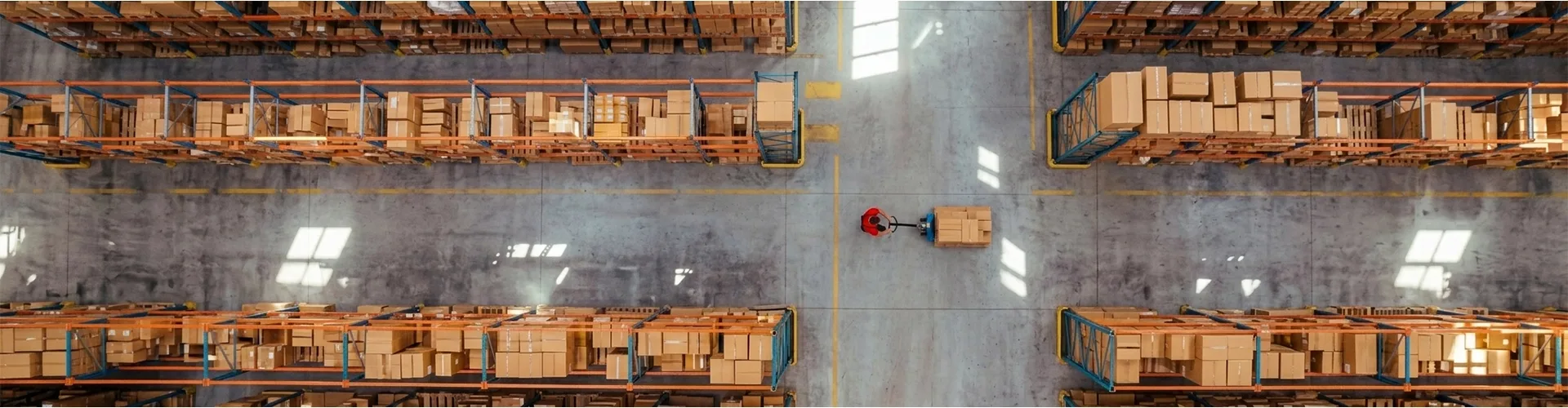 An overhead view of a warehouse worker using a pallet jack to move a box through an aisle between tall storage racks filled with cardboard boxes.