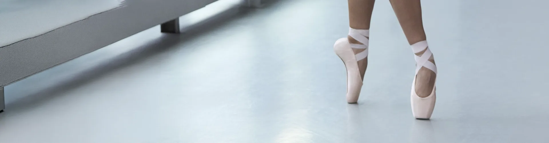A ballet dancer stands on tiptoe in pink pointe shoes on a smooth studio floor near a bench.