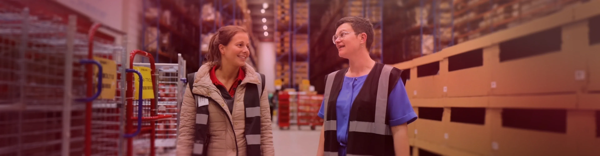 Two women wearing reflective vests walk and talk in a warehouse aisle lined with shelves and storage bins.