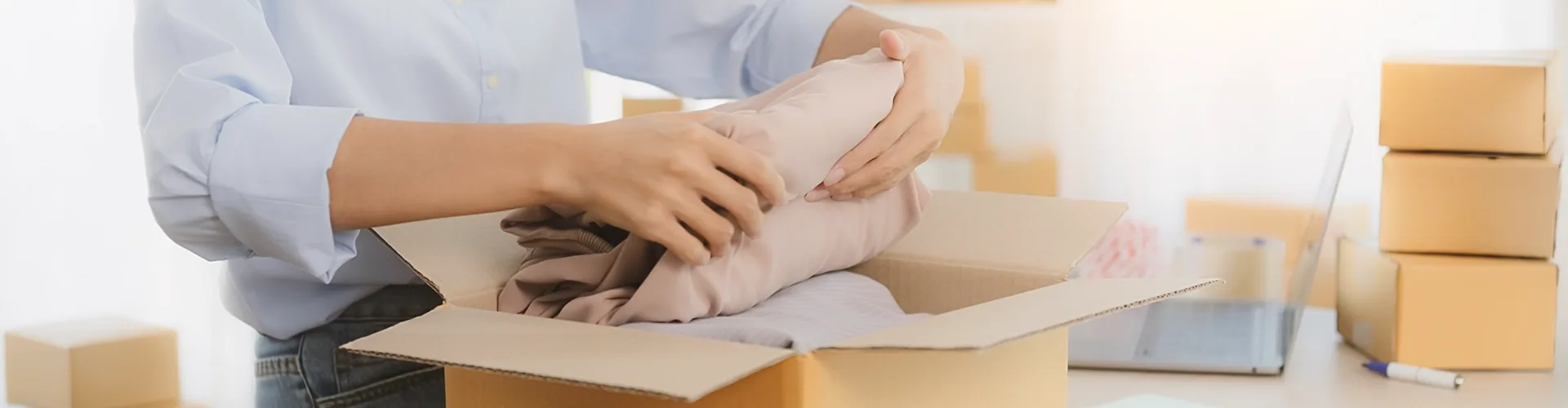 A person places folded clothes into an open cardboard box, with several closed boxes and a laptop visible in the background.