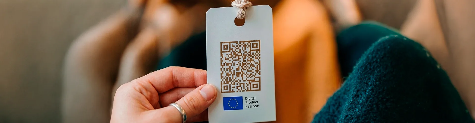 A hand holds a product tag with a QR code and an EU Digital Product Passport logo; blurred background with a person sitting.