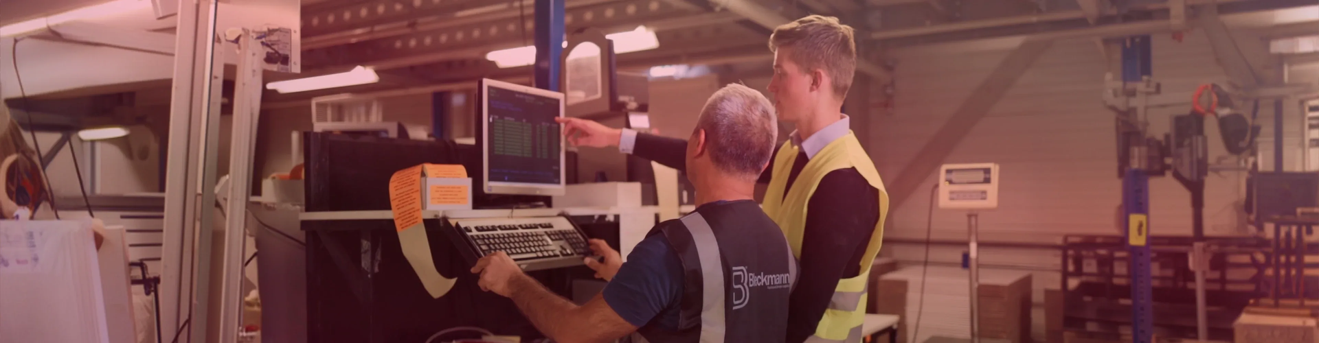 Two men, one in a safety vest, stand in a factory while reviewing data on a computer screen next to industrial equipment.