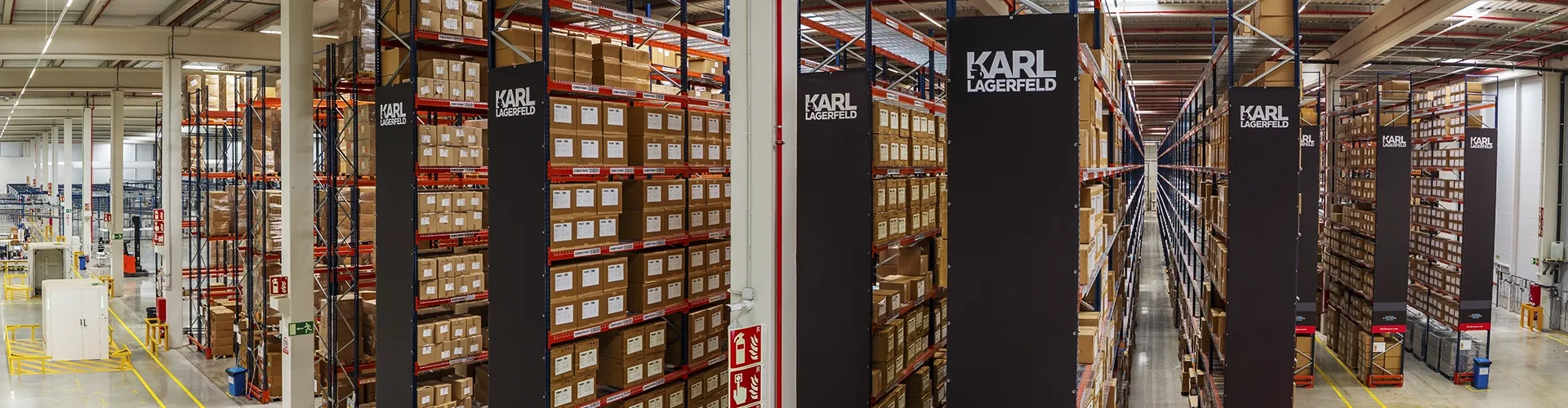 A large warehouse with tall shelves stacked with boxes labeled "Karl Lagerfeld" under bright overhead lighting.