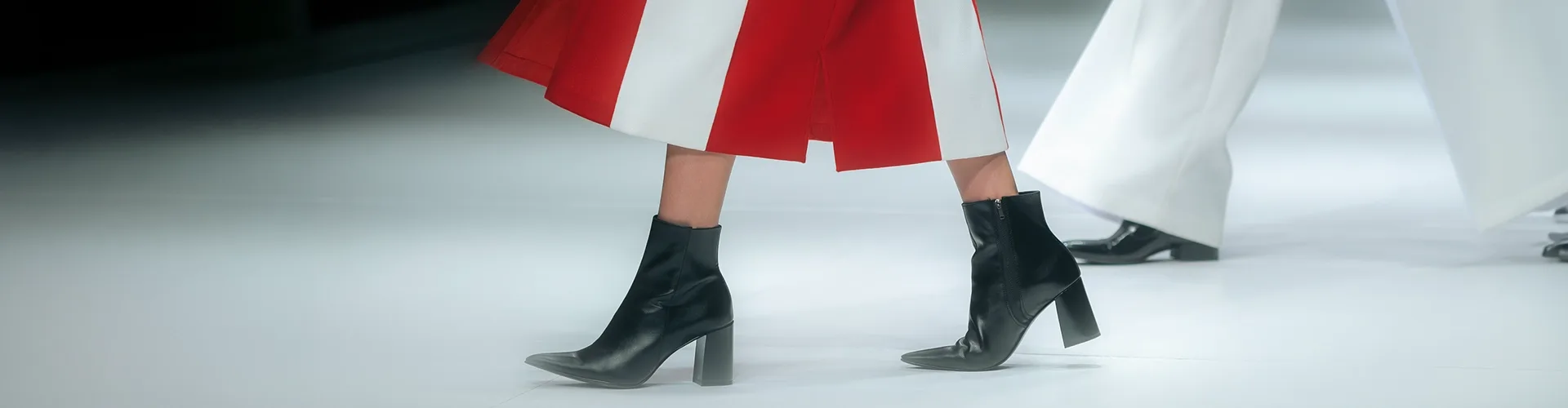 Two people walking on a runway; one wears a red and white skirt with black heeled ankle boots, the other wears white pants with matching white footwear.