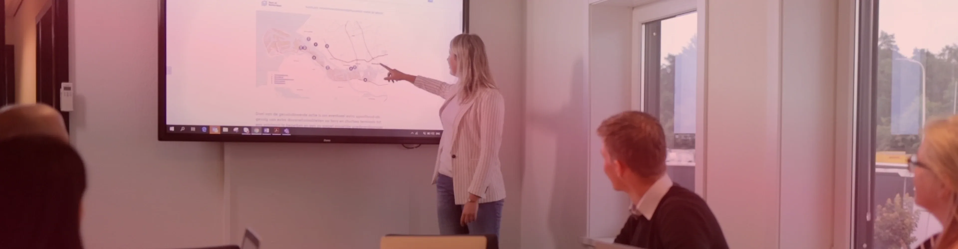 A woman stands and points at a map on a large screen while two people seated at a conference table look on.