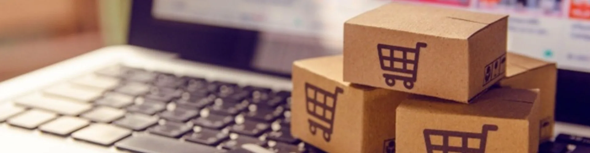 Small cardboard boxes with shopping cart icons are stacked on a laptop keyboard, with a blurred online shopping website visible on the screen in the background.