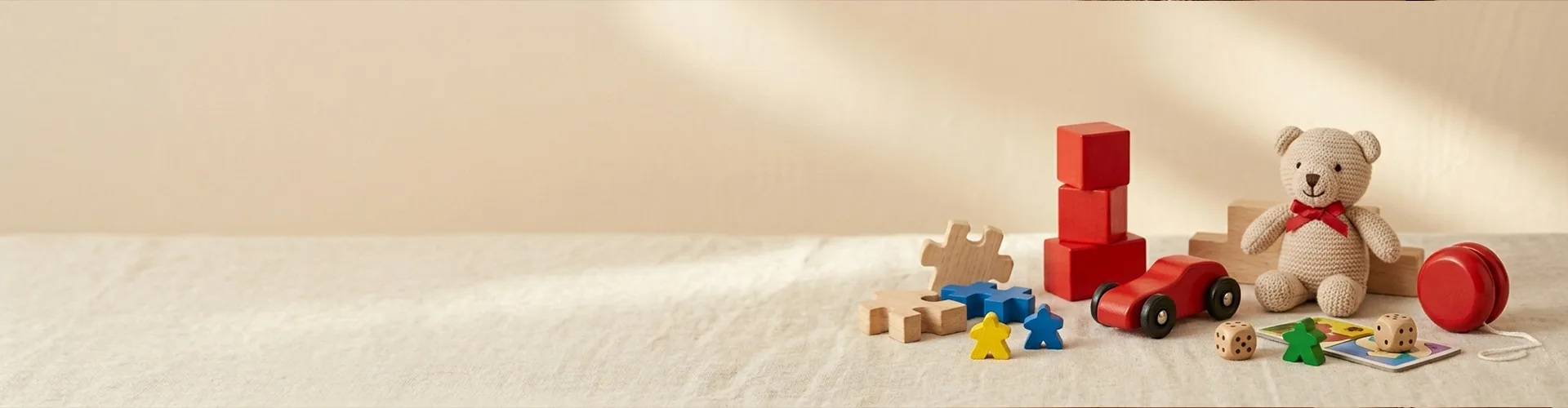 A group of children's toys including a teddy bear, red toy car, wooden blocks, puzzle pieces, dice, a red yo-yo, and colored game pieces arranged on a light surface.