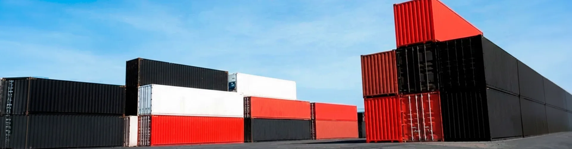 Stacks of shipping containers in black, white, and red are arranged on a paved lot under a clear blue sky.