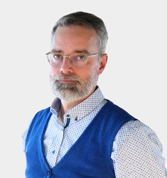 A man with a beard and glasses is wearing a blue vest over a patterned shirt, standing against a plain background.