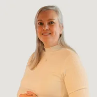 A woman with long gray hair wearing a white ribbed turtleneck top stands against a plain background, smiling at the camera.