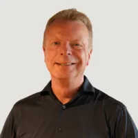 A middle-aged man with short light hair is smiling, wearing a dark collared shirt, standing against a plain light background.