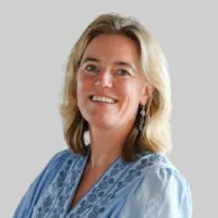 A woman with blonde hair wearing a light blue patterned blouse and earrings smiles at the camera against a plain light background.