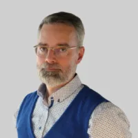 A middle-aged man with glasses, short gray hair, and a beard, wearing a patterned shirt and blue vest, standing against a plain light background.