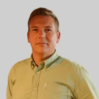 A man with short blond hair wearing a light green button-up shirt stands against a plain light gray background.