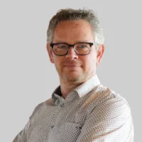 A man with short curly hair, glasses, and a beard is wearing a patterned button-up shirt and standing against a plain light gray background.