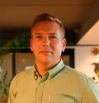 A man with short blond hair wears a light green button-up shirt and stands indoors with soft lighting and blurred background.